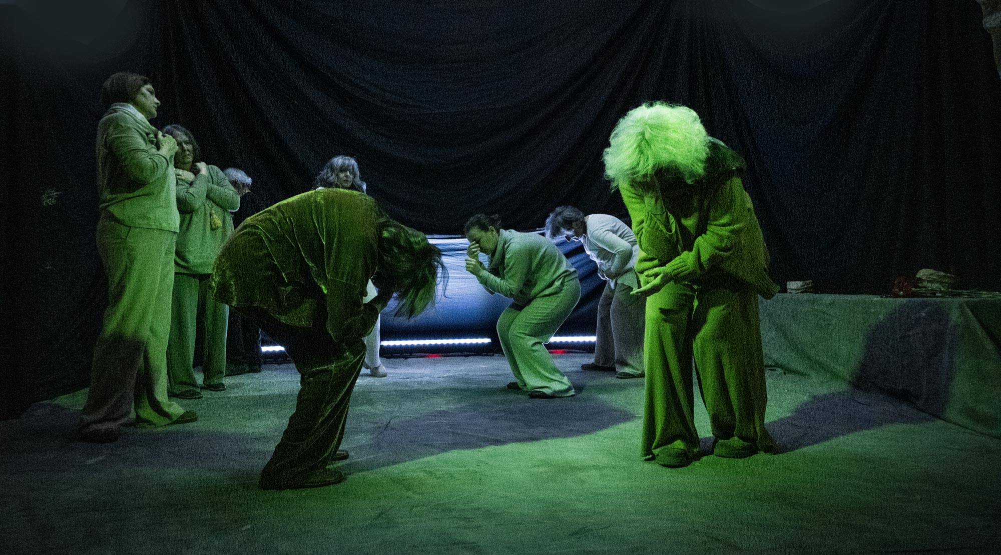 Gruppo di attori e attrici in un momento di rappresentazione di disperazione collettiva inondati da una luce verde.